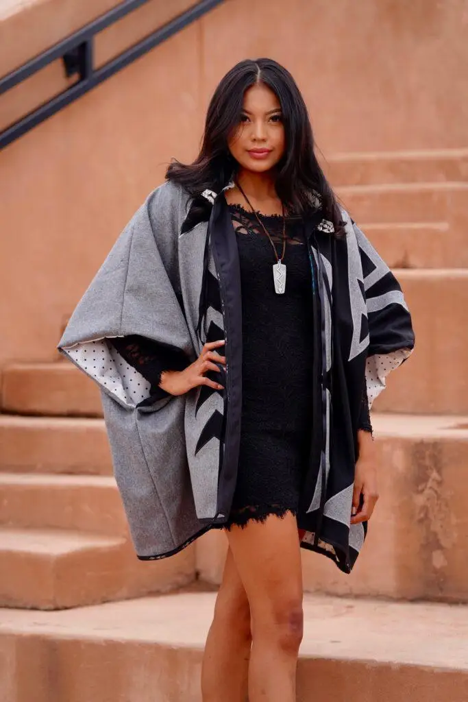 native american woman with hands on hips wearing gray jacket with large billowing sleeves, a dark dress underneath, standing in front of terra cotta stairs