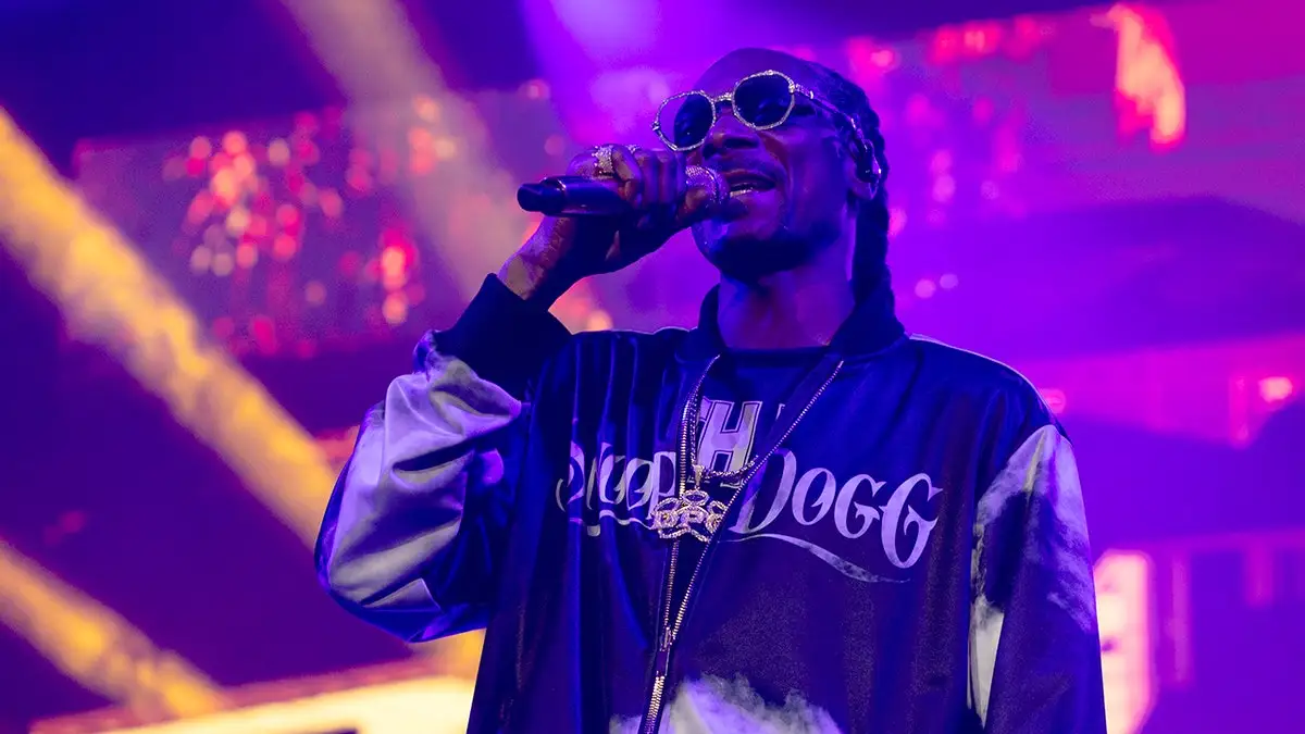 Snoop Dogg in a jacket that says his name performs on stage wearing sunglasses