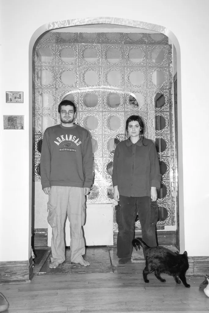 This black and white photo shows the gallerists standing in a doorway with a clear patterned wall behind them. A black cat walks in the foreground. Davis wears an Arkansas sweatshirt and cargo pants while Harvard wears baggy pants and a buttonup shirt.