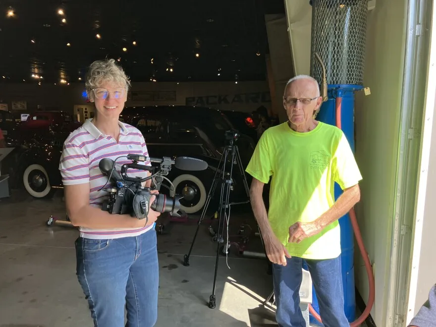 A woman holding a camera poses with a man outside a car museum.