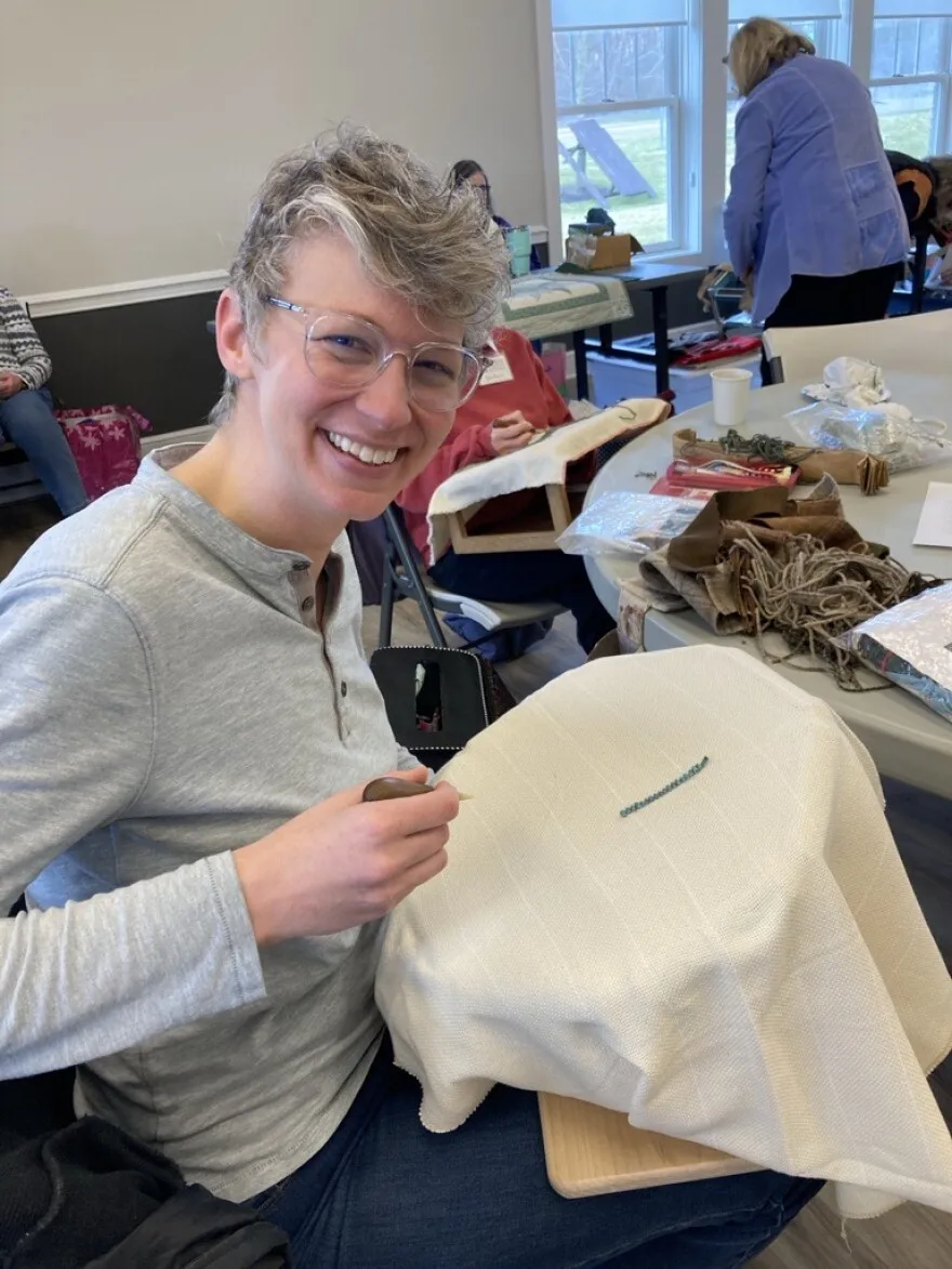 A seated woman stitches fabric through a linen canvas.