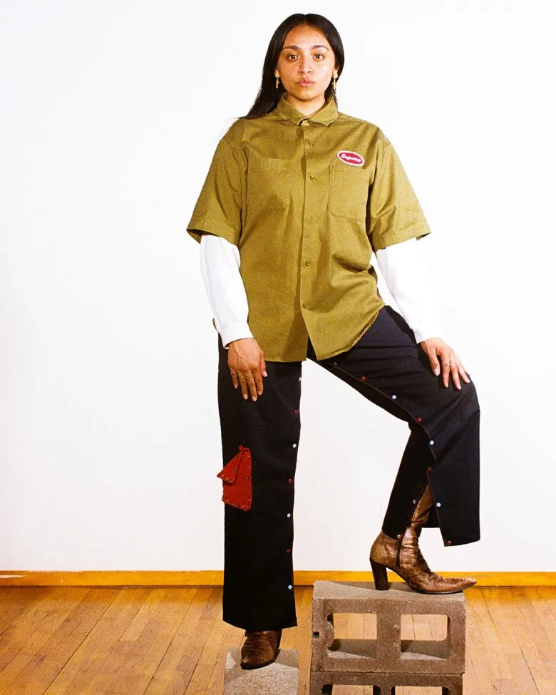 Delgado wears button-up black pants and a khaki workshirt over a long-sleeved white shirt. She has on brown cowboy boots. She wears her black hair parted down the middle and stands facing the camera. A white wall is behind her. One foot is propped up on a couple cinder blocks, the other stands on one cinder block.