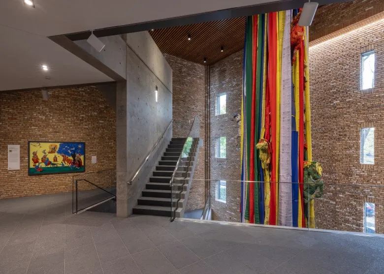 Horizontal view of the interior brick atrium of Wrightwood 659. To the right, in a floors-long opening, hang a thick mass of prayer flags, in green, white, orange, yellow and blue. To the left are wall-mounted works.