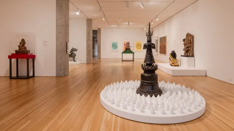 Installation view of Reimagine shows various ancient artifacts, including sculptures of deities in metals and stone on various colored plinths throughout the gallery. In the background of various wall-hung prints and paintings.