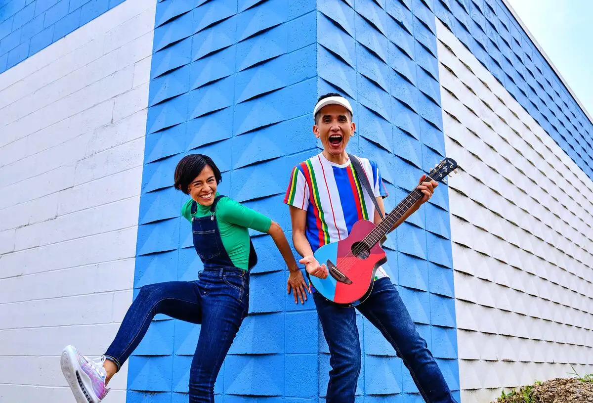 Two young people in jeans and colorful shirts, one with a guitar, in front of a blue and white building