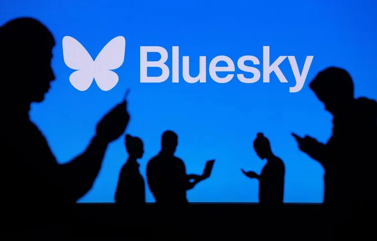 The silhouettes of several people using digital devices against the blue backdrop of the Bluesky logo.