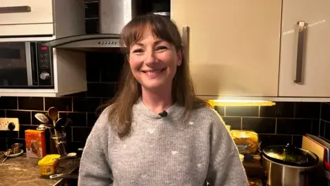 Colette, with brown hair and wearing a grey jumper smiles at the camera. She is standing in front of a black kitchen hob.