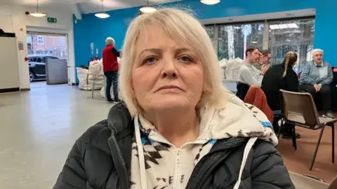 Julie, with blonde hair looks at the camera. She is wearing a zip up fleece with a black and blue patter and a black puffy coat. People are sitting on chairs behind her.