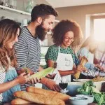 Good friends laughing and talking while preparing meals at table full of vegetables and pasta ready for cooking in kitchen