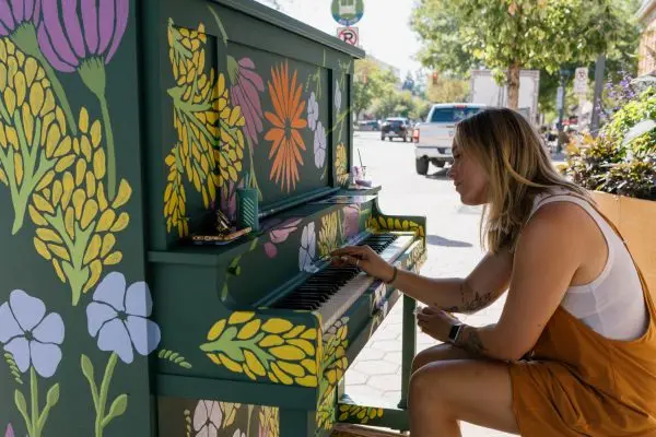 A woman delicately paints a green piano with colorful yellow, purple, and orange flowers.