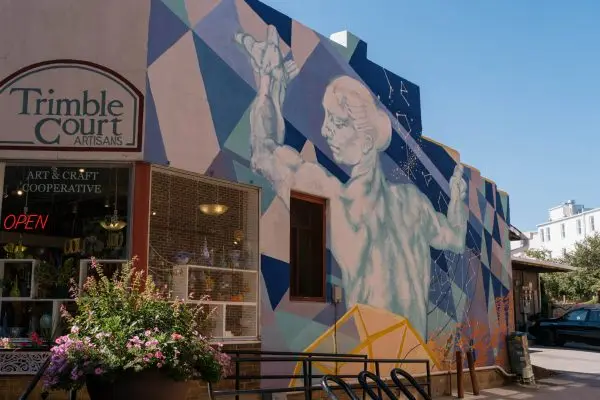 A mural painted on the wall of an alleyway building depicts a marble statue on a colorful background.