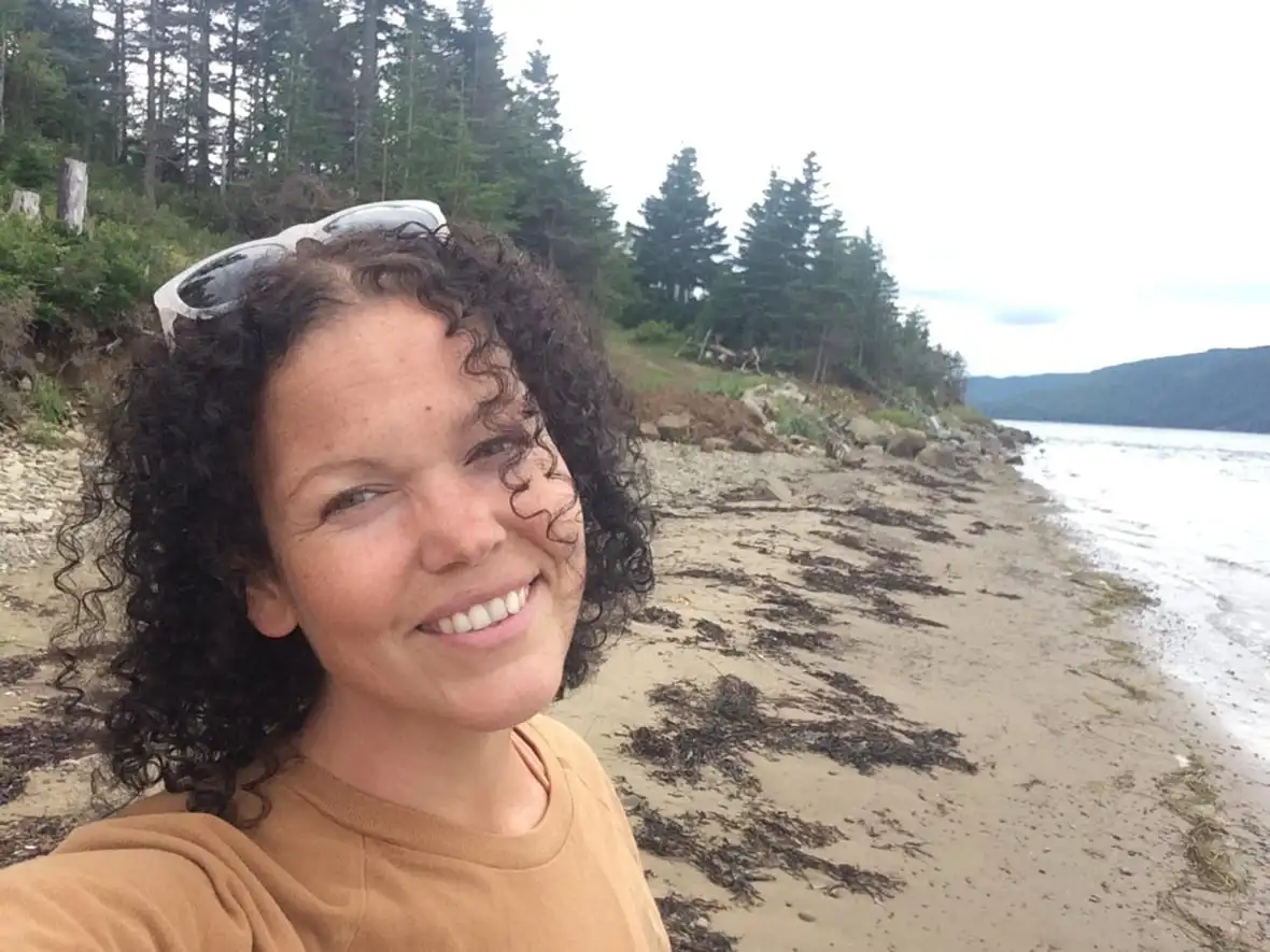 woman with dark curly hair and white sunglasses on her head smiles into camera while standing on a beach.