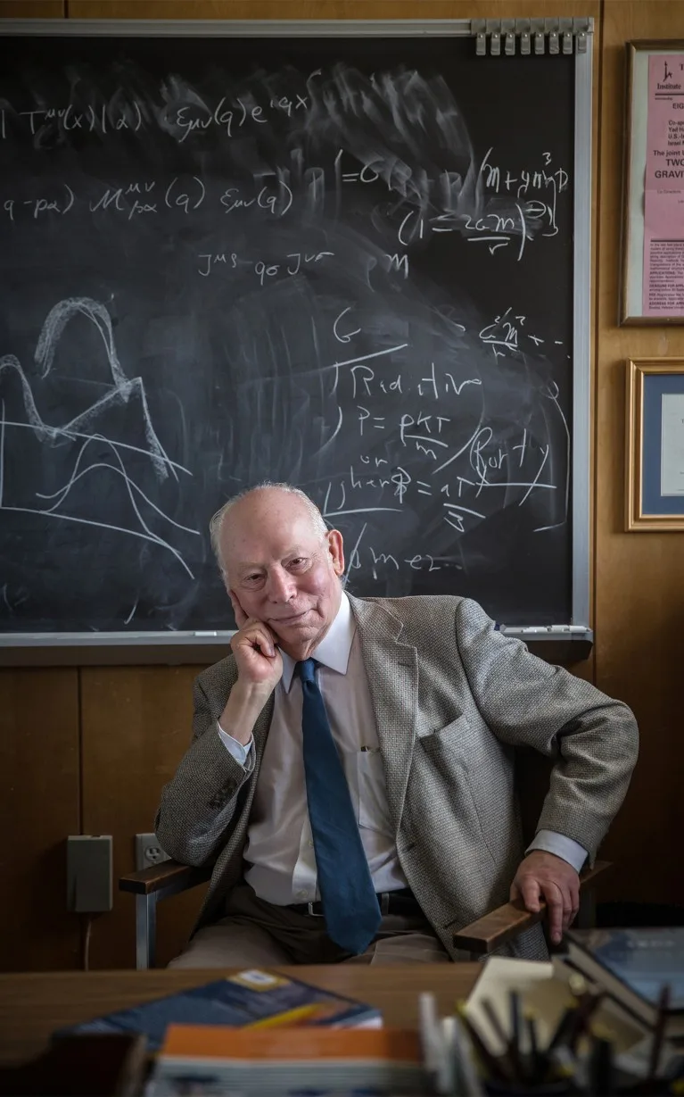 Steven Weinberg sits at a desk on front of a backboard full of equations