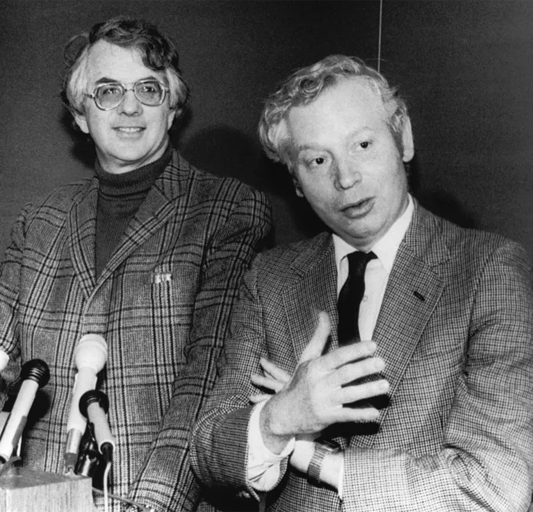 Sheldon Glashow and Steven Weinberg stand behind some microphones