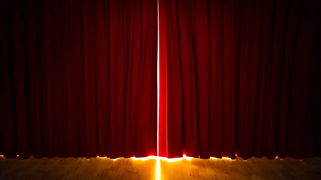 A bright light peeking through a set of closed, red theater curtains.