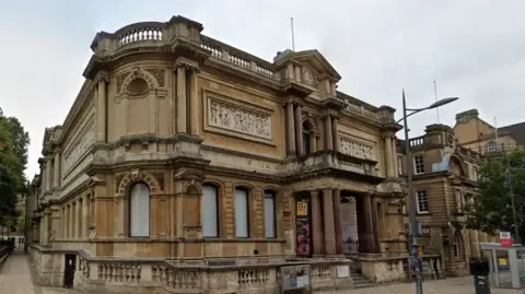 Google Wolverhampton Art gallery is a brown-beige stone building with people carved into a design near the roof. There are pillars around the entrance which has a small flight of steps. There is also a balcony above the entrance which also has brown pillars on it.