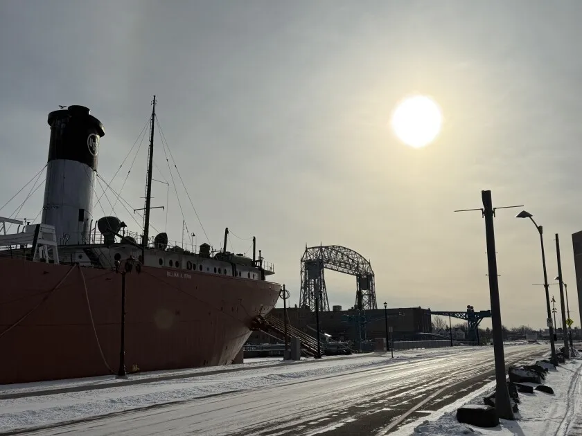 View of icy road with stern of bulk cargo freighter at left, with lift bridge in background. Sun shines through hazy morning sky.