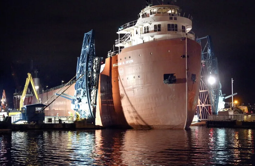 Bulk cargo freighter passes narrowly through lifted halves of a blue drawbridge at night, with bright lights reflected in water surface.