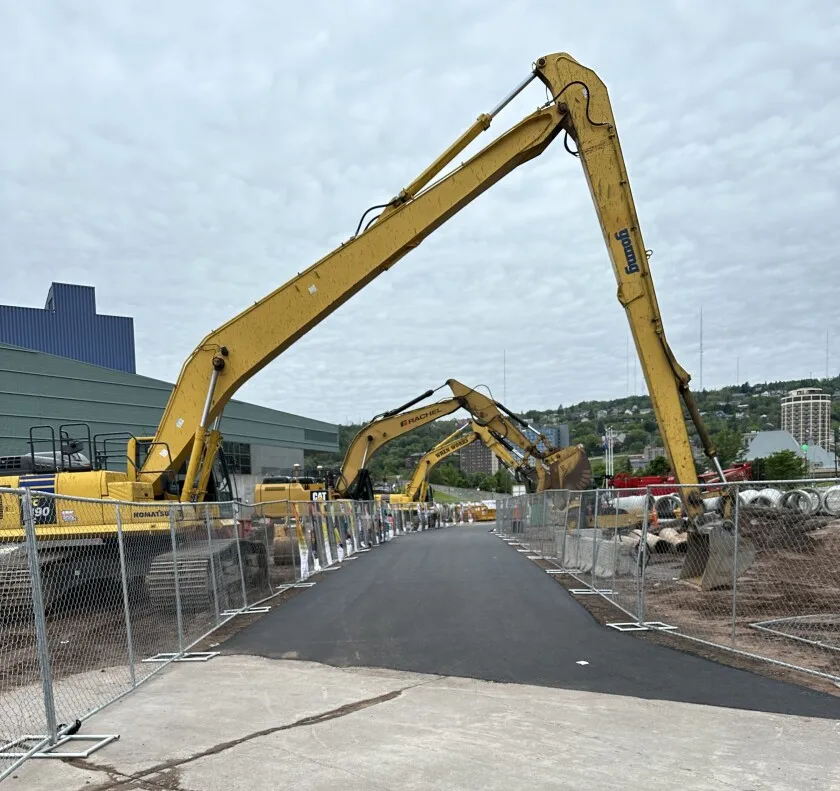 Three yellow excavator arms arc over paved pathway, with large blocky building at left and urban hillside in background.