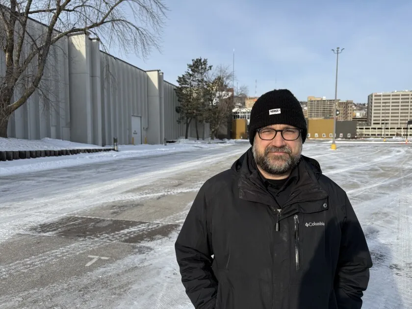 White man wearing black winter garb stands in empty parking lot in front of blocky white building, with hillside cityscape in background.