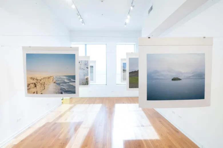 Two rows of large landscape photographs by Linye Jiang hang from wires across the gallery. Large windows are in the back, which stream bright sunlight across the room. In the foreground on the right is a photo of a vast body of water with a rock in the center; on the left is a rocky coast beside a sea.