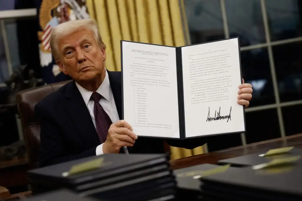 a man with a fake tan in a suit holds up a piece of paper with his signature on it while seated at a large desk