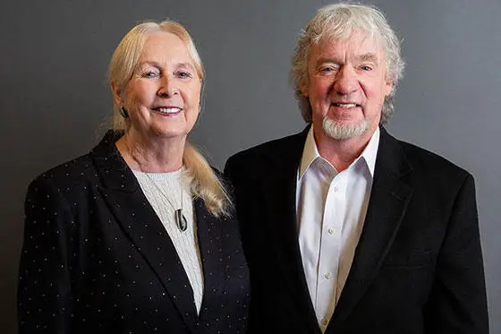 Image of a light-haired woman with smile on left and grey-haired man with smile on right. Both are wearing dark jackets.