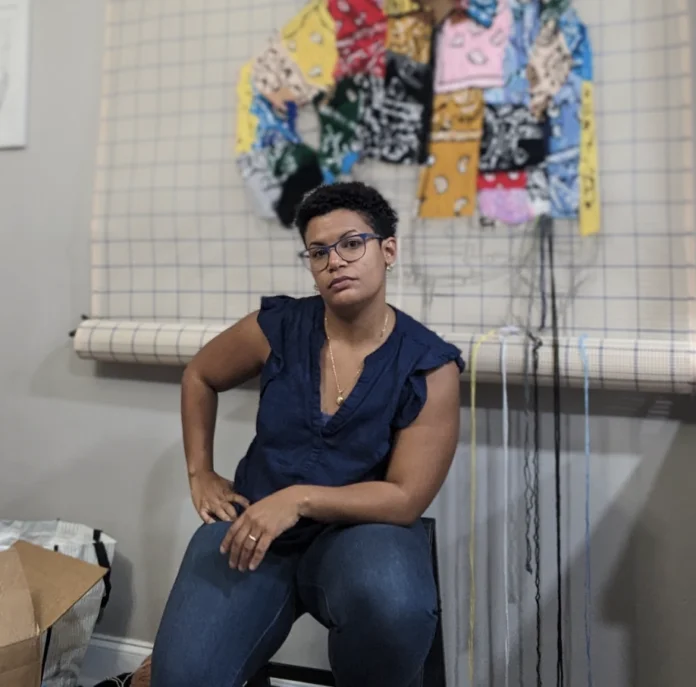 Person sitting on a stool in front of woven textile art. They are wearing glasses and a blue shirt.