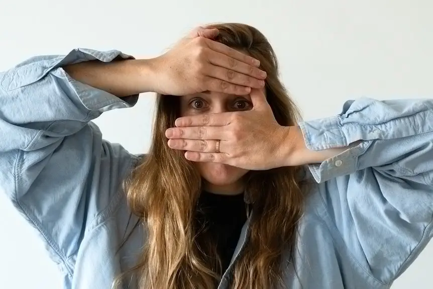 CJ Hendry, with long fair hair and blue shirt, holds two hands over her face with only eyes visible.