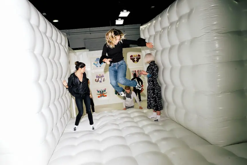 Three people jumping between soft inflated white walls and a white floor, like a bouncy castle, smiling and laughing.