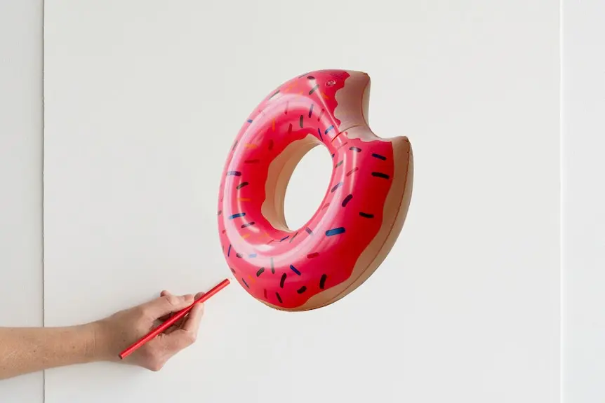 Hand holds pencil near drawing on white canvas of bright pink doughnut shaped inflatable pool ring.