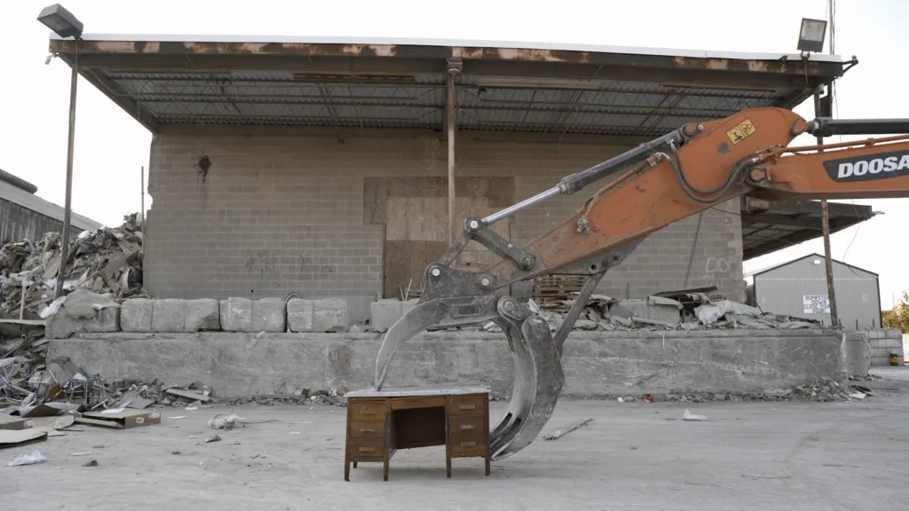 six drawer desk about to be picked up by a bulldozer claw in front of a cinderblock building