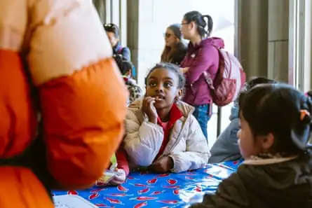 A child listens to an adult while putting her hand near her mouth