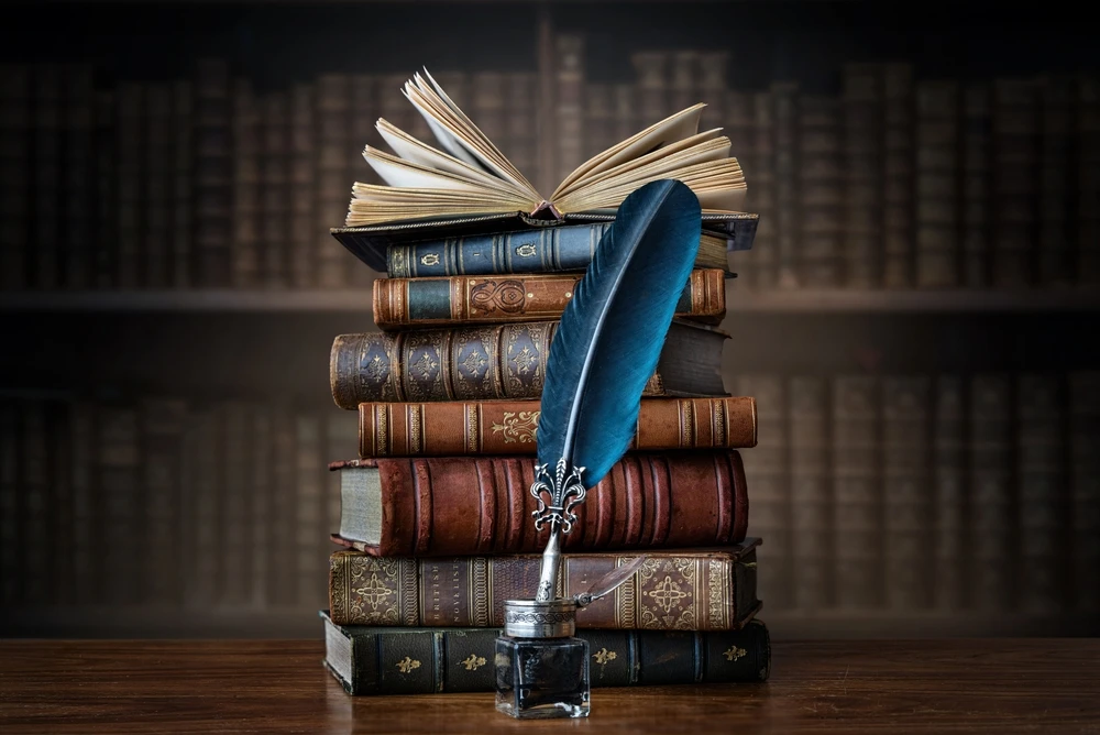 Old books, quill pen, and vintage inkwell on wooden desk in old library.