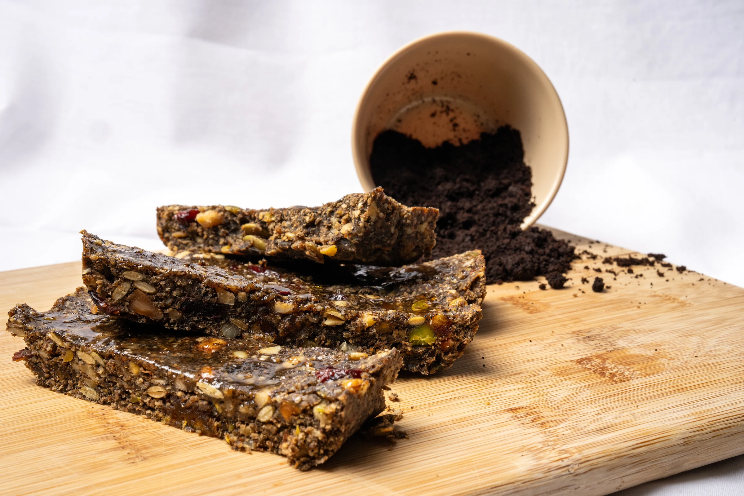 A close-up of rectangular protein bars placed on a wooden cutting board along with a cup of coffee grounds tipped over in the background.