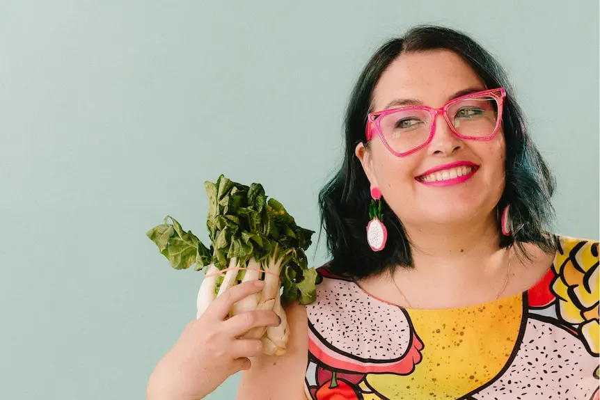 A woman with shoulder-length black hair, in fruit-patterned dress and pink-framed glasses, holding a bunch of leafy greens.