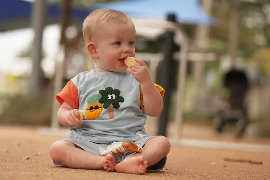 A young child sitting on the floor at a playground eating a cracker.