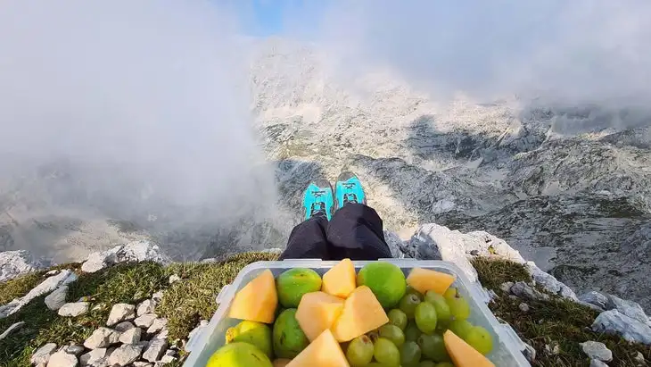 Image of hiker's feet in boots, with a container of fruit in the foreground and a mountain vista behind.