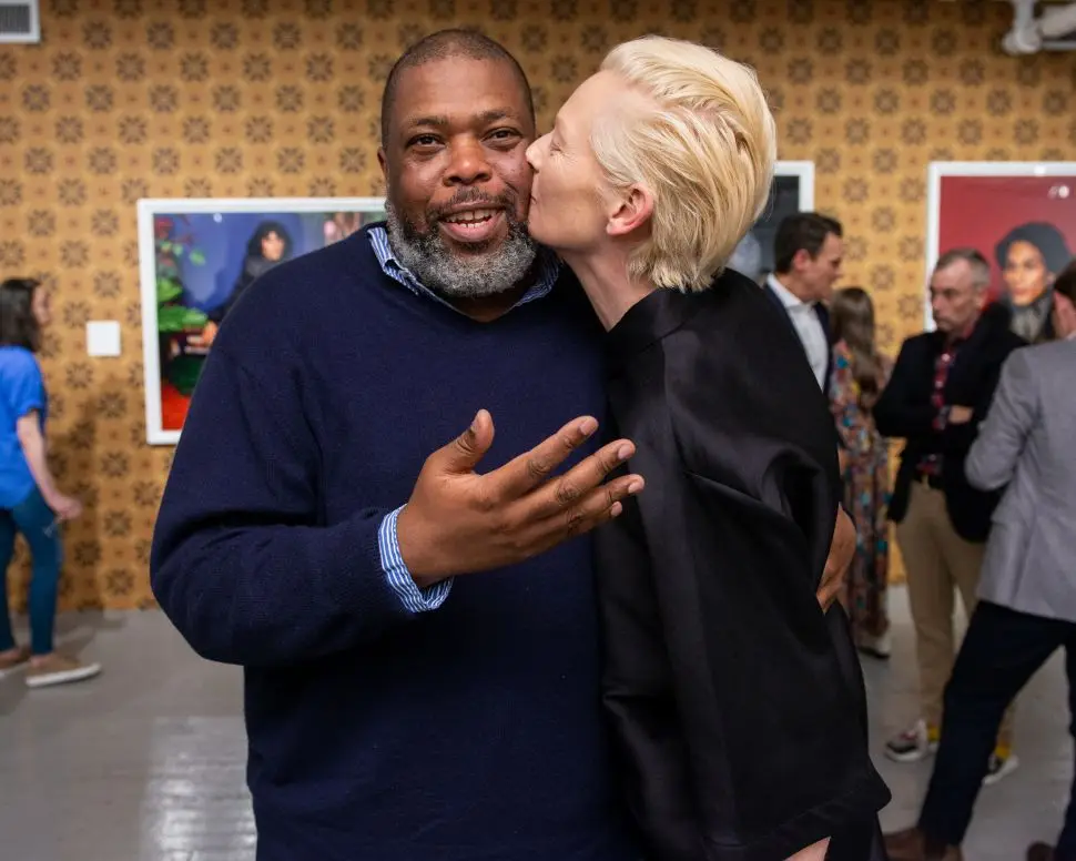 A woman with slicked back blonde hair kisses a sweater-wearing man on the cheek as he gazes into the camera