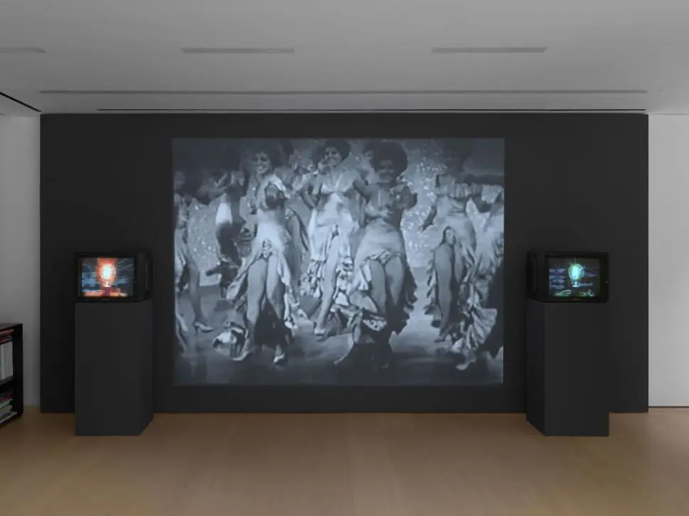 A video installation features a large black-and-white projection of dancers in vintage costumes, flanked by two small screens showing abstract glowing images, all displayed on dark pedestals.