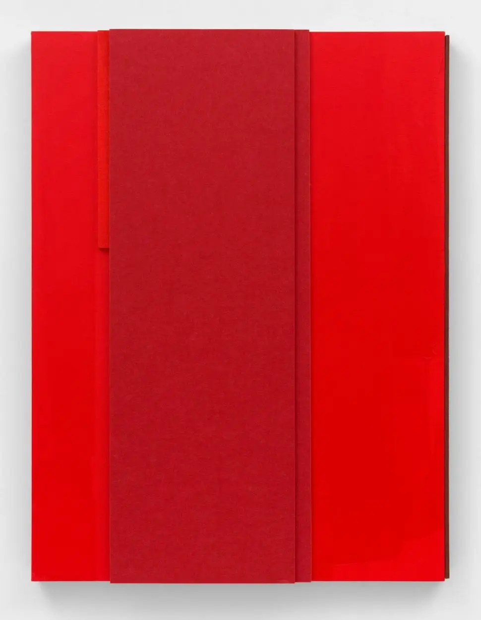 A modern abstract artwork with overlapping rectangular panels in shades of red is mounted on a white wall, emphasizing texture and geometric layering.