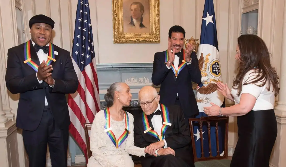 Five people in black tie dress laugh and talk in a room while wearing rainbow ribbons signifying their designation at Kennedy Center honorees. 