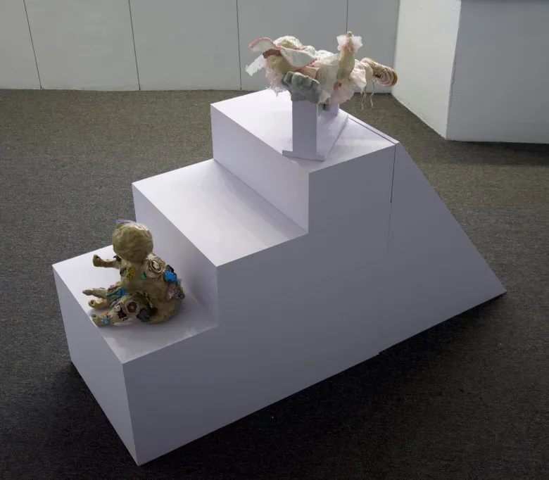 Two baby sculptures are installed on a set of steps made out of wood and painted lavender installed in the center of the gallery. The image is taken from above. One baby is on the bottom step, seated with its hands extending out, and the other is installed in two large hands on the top step.