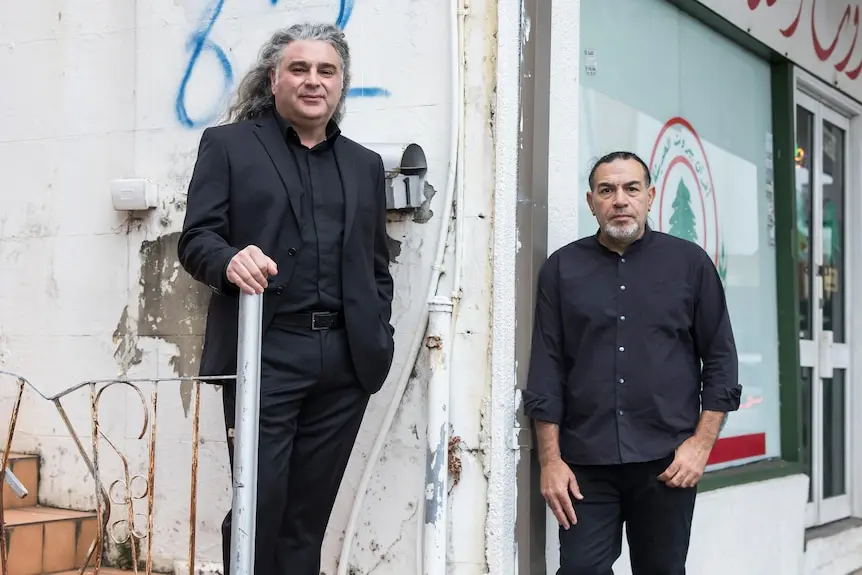 Two man stand on street wearing black suits. One smiles and one has serious expression.