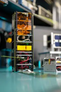 A boxy satellite that's 30 cm tall by 10 cm wide and 10 cm deep sits on a workbench. Two smaller CubeSats, each 10 cm cubes, can be seen in the background.