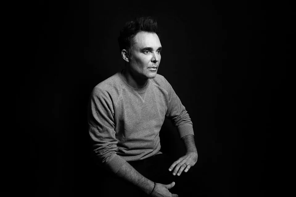 A person seated against a dark background, wearing a gray sweater, with hands resting on their knee, exuding a contemplative mood.