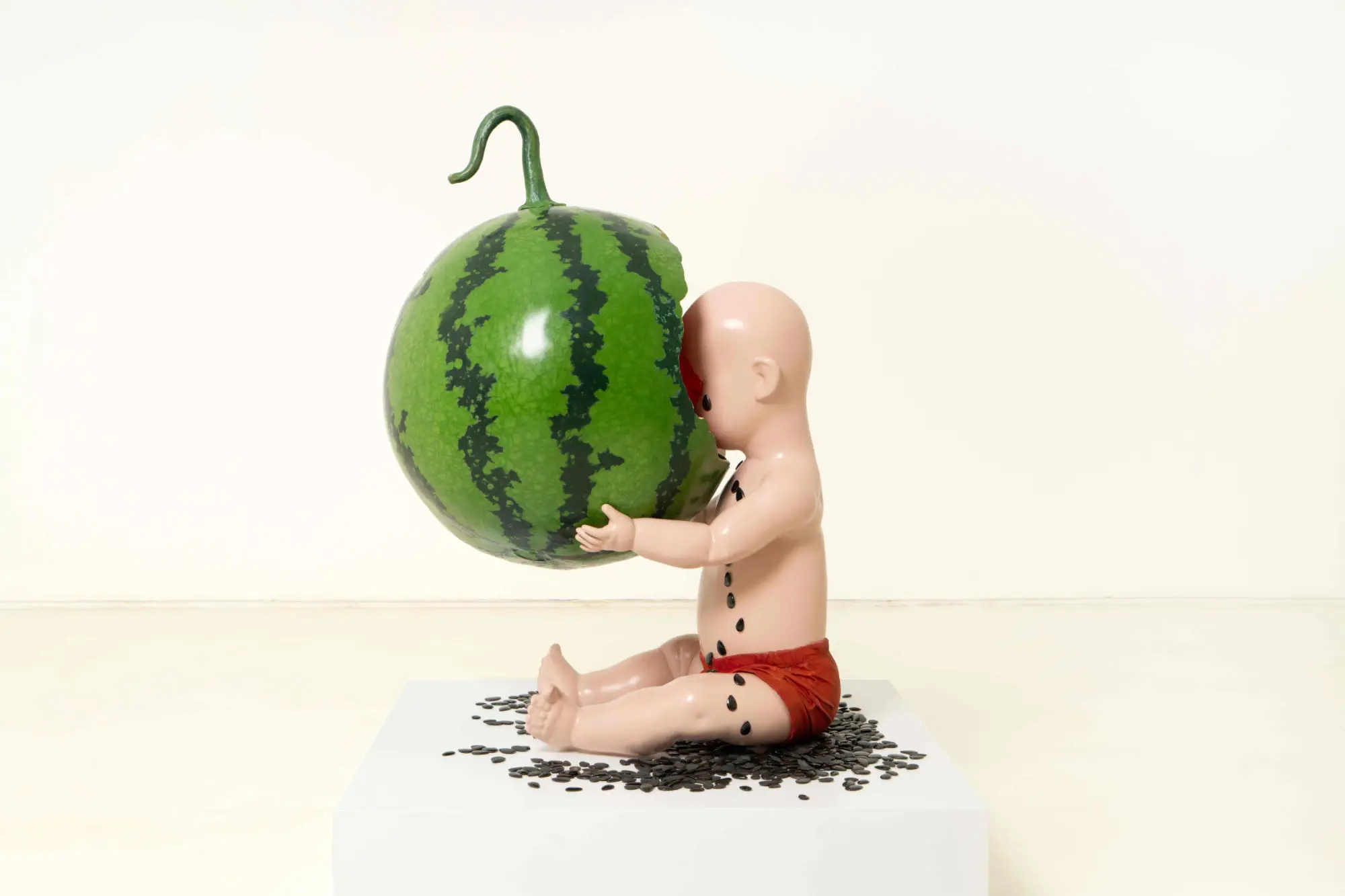 Renee Jin sculpture of a baby seen in profile devouring an oversized watermelon with he seeds spilling onto the child and the floor. 