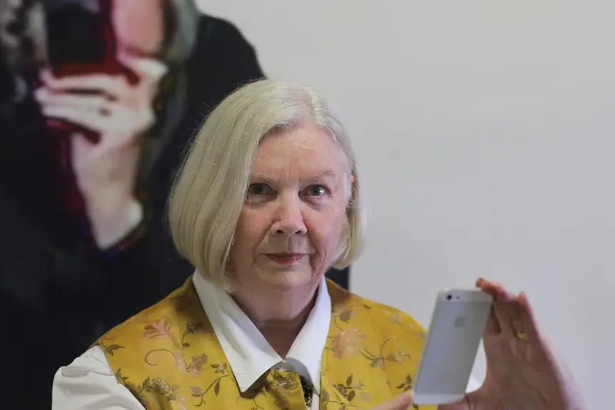 Greer Honeywill, in yellow vest and smiling with closed mouth, holds phone. In background is photo of her holding a phone.