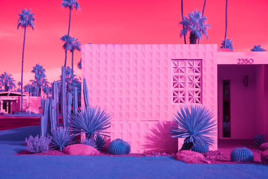 Bright pink, blue and purple image of a house surrounded by cacti, scrubs and palm trees.