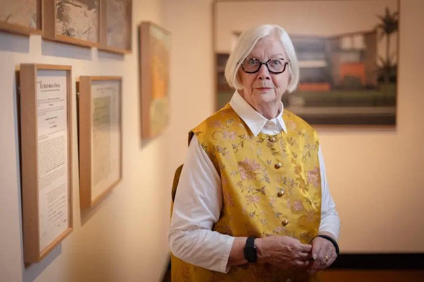 A woman with grey hair wearing a yellow vest standing in an art exhibition.